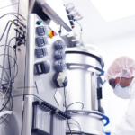 Worker in protective clothing beside stainless steel bioprocess equipment and a control panel.